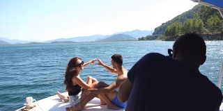 一对年轻的情侣坐在船头，对着不认识的摄影师摆姿势。幸福的情侣在船上共度时光，享受夏天的旅行。假期的概念。