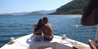 一对不认识的年轻夫妇坐在船头，对着不认识的摄影师摆姿势。幸福的情侣在船上共度时光，享受夏天的旅行。假期的概念。后方后视图