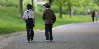 学龄前的小男孩，可爱的孩子，兄弟，穿着复古风格的衣服，在公园里盛开的树下，看起来时髦，走在一条小路上