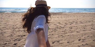 一个面目全非的女孩牵着一个男人的手在海滩上奔向大海。跟着我拍的年轻女子戴着帽子拉她的男朋友在海边。暑假或假日概念。观点慢动作