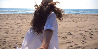 一个面目全非的女孩牵着一个男人的手在海滩上奔向大海。跟着我拍的年轻女子戴着帽子拉她的男朋友在海边。暑假或假日概念。观点慢动作