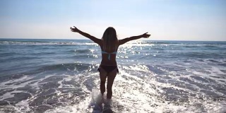 一名年轻女子走在海边，举手示意。一个无法辨认的美丽女孩在海边享受着生活和自由。暑假或假日概念。以自然景观为背景。慢镜头后视图