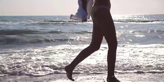 日出时，穿着比基尼和衬衫的陌生女人的脚在海浪上奔跑。年轻美丽的女孩慢跑和在海边玩。暑假或假日概念。关闭侧视图慢动作