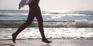 日出时，穿着比基尼和衬衫的陌生女人的脚在海浪上奔跑。年轻美丽的女孩慢跑和在海边玩。暑假或假日概念。关闭侧视图慢动作