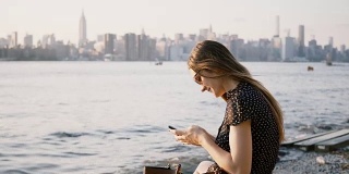 迷人的白人女孩戴着墨镜使用智能手机messenger应用程序，在纽约河滩周围看4K