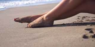 美丽的女性的腿躺在沙滩上，放松在海边。在阳光明媚的日子里，一个面目全非的年轻女子坐在海边晒太阳。度假胜地女孩的脚。暑假或假日概念。关闭侧视图慢动作