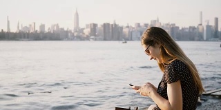 美丽的欧洲女孩与长头发太阳镜使用智能手机购物网上应用程序在纽约日落海滩4K