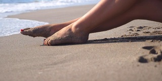 美丽的女性的腿躺在沙滩上，放松在海边。在阳光明媚的日子里，一个面目全非的年轻女子坐在海边晒太阳。度假胜地女孩的脚。暑假或假日概念。关闭侧视图慢动作