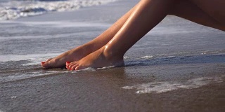 海浪冲刷着女性晒黑的脚。性感女孩的腿躺在海边的金色沙滩上，放松在暑假旅行。旅行的概念。慢动作关闭低角度视图侧视图