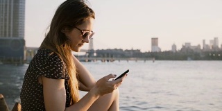 体贴的白人女孩在太阳镜使用智能手机社交网络应用程序，享受惊人的日落在城市海滩4K