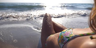 难以辨认的性感女孩躺在海滩的波浪中。美丽的年轻女子放松在海边的暑假旅行。海浪冲刷着晒黑的女性身体。慢动作特写后视图