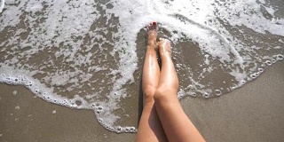 海浪冲刷着晒黑的女性脚。在一个阳光明媚的日子里，一个美丽的女孩躺在沙滩上。暑假期间，一个陌生的年轻女子在海边放松。俯视图慢动作特写