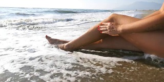 海浪冲刷着年轻女性的脚。难以辨认的迷人的女人在比基尼躺在海滩上。晒黑的女孩在美丽的海岸晒太阳。暑假或假日概念。关闭慢动作侧视图低角度视图