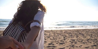 年轻的女孩牵着男人的手，在热带异国的海滩上奔向大海。跟着我拍的一个年轻女人拉着她的男朋友在海边。暑假或假日概念。观点慢动作