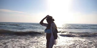 年轻女孩穿着比基尼和衬衫，背包在海边的海滩上旋转。戴着帽子的漂亮女人，在阳光灿烂的日子里享受生活，在海边玩得开心。暑假或假日概念。特写慢动作