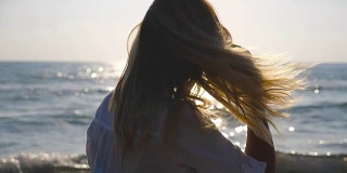 年轻女孩沿着海岸散步，玩弄着她的金发。美丽的女人在暑假旅行中享受生活。背景是平静的海浪。关闭侧视图慢动作