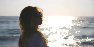 年轻的女孩穿着衬衫沿着海岸走着，抚摸着她的金发。戴着太阳镜的美女在暑假旅行中享受生活。背景是平静的海浪。特写慢动作侧视图