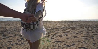 跟着我拍的年轻女子穿着衬衫背着背包拉着男友去海边。美丽的女孩牵着男人的手在空旷的沙滩上奔跑。暑假或假期的概念。慢镜头观点