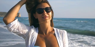 美丽的女孩戴着太阳镜，微笑着玩她的金发在海岸。晒黑的女性享受生活和在海边玩。暑假或假期的概念。慢镜头