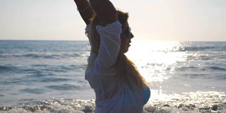 穿着比基尼和衬衫的年轻女子在海岸上旋转，并举起她的手。美丽的金发女孩沿着海边散步，享受生活。暑假放松一下。特写慢动作侧视图