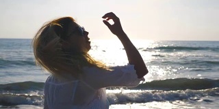 侧面美丽的女孩在比基尼和衬衫走在海岸和触摸她的金发。年轻女子在海滩上享受暑假。海边的背景。关闭侧视图慢动作
