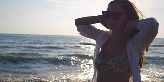 快乐的女人在阳光明媚的海滩上散步和旋转。年轻的女孩戴着太阳镜，微笑着玩弄她的头发。女士在旅行中享受生活。暑假或假期。慢镜头