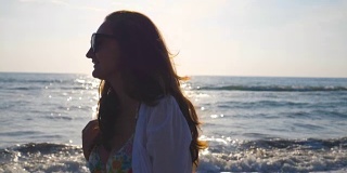 侧面美丽的女孩在比基尼和衬衫走在海边和玩她的头发。快乐的年轻女人戴着太阳镜，在暑假里享受生活。慢动作特写侧视图