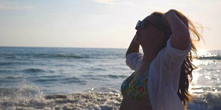 侧面美丽的女孩在比基尼和衬衫走在海边和玩她的头发。快乐的年轻女人戴着太阳镜，在暑假里享受生活。慢动作特写侧视图