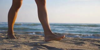 女性的脚踩在沙滩上的特写与海浪的背景。一个美丽的女人在阳光明媚的日子赤脚走在海边。暑假或假期的概念。慢动作低角度视图