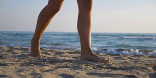 在阳光明媚的日子里，在海滩上赤脚行走的女性脚的低角度视图。年轻女子的双腿踩在沙子上。赤脚女孩在海边。波在背景。暑假或假日概念。特写慢动作