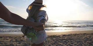 跟着我拍的年轻女子在衬衫与背包拉她的男友到岸边。美丽的女孩戴着帽子牵着男人的手，在空旷的海滩上向大海跑去。暑假的概念。慢镜头观点