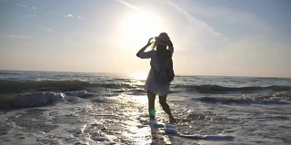 快乐的女孩穿着比基尼和衬衫，背包慢跑到海边，用她的脚泼水。戴着帽子的漂亮女人享受着自由和户外的乐趣。夏季旅游的概念。后视图慢动作