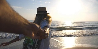 美丽的女孩在衬衫与背包牵着男性的手，在空旷的海滩上奔跑。跟着我拍的快乐女人戴着帽子拉着男友去海边。暑假的概念。慢镜头观点