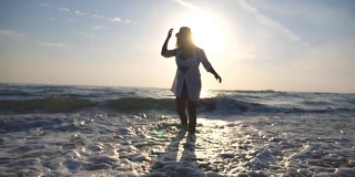 快乐的年轻女孩穿着比基尼和衬衫，背包慢跑到海边，用她的脚泼水。美丽的女人在海边旋转，享受户外的乐趣。暑假的概念。慢动作