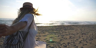小女孩穿着衬衫，背着双肩包牵着男人的手，带着她去了海边。跟着我拍的幸福美丽的女人戴着帽子拉着男友去海边。情侣在一起度假。夏天旅游的概念。慢