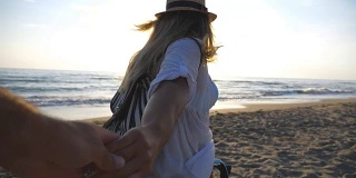 跟着我拍的年轻女子穿着衬衫背着背包拉着男友去海边。漂亮的女孩戴着帽子牵着男人的手在空旷的海滩上奔跑。幸福的夫妇花时间在一起。慢镜头观点
