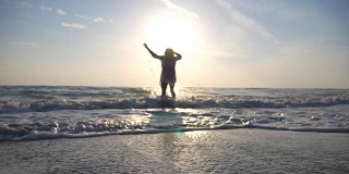 年轻快乐的女人穿着衬衫和背包跑到海边，用她的脚泼水在日出。漂亮的女孩戴着帽子享受暑假或假期。旅行的概念。后视图慢动作