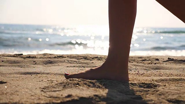 近距离的女性的脚走在金色的沙滩与海浪的背景。年轻女子的双腿踩在沙子上。光着脚的女孩在海边。暑假或假日概念。慢动作低角度视图侧视图