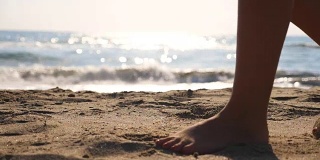 近距离的女性的脚走在金色的沙滩与海浪的背景。年轻女子的双腿踩在沙子上。光着脚的女孩在海边。暑假或假日概念。慢动作低角度视图侧视图