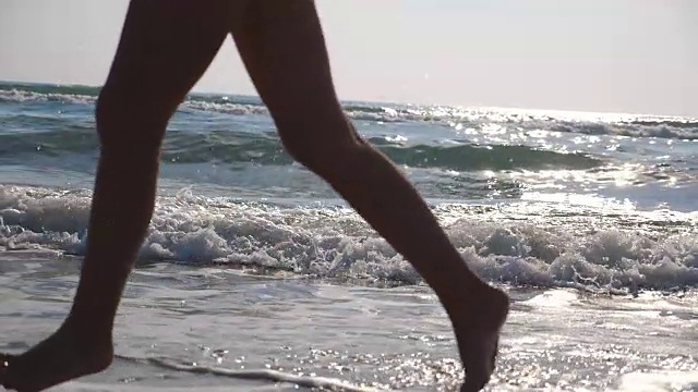 女性的脚在潮湿的沙子和海浪在海滩的海水上奔跑的特写。年轻女子在岸边奔跑的双腿。光着脚的女孩在海边。暑假或假日概念。慢动作侧视图低角度视图