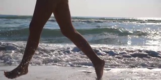 女性的脚在潮湿的沙子和海浪在海滩的海水上奔跑的特写。年轻女子在岸边奔跑的双腿。光着脚的女孩在海边。暑假或假日概念。慢动作侧视图低角度视图