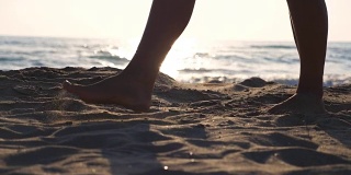 近距离的女性的脚走在金色的沙滩与海浪的背景。年轻女子的双腿踩在沙子上。光着脚的女孩在海边。暑假或假日概念。慢动作低角度视图侧视图