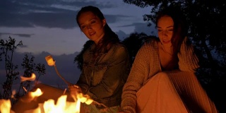 两个年轻的白人女孩晚上坐在篝火旁，准备一个棉花糖，看着篝火，思考着
