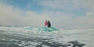 年轻的夫妇有乐趣在冬季散步的背景冰冻结的湖。情侣们坐在巨大的蓝色浮冰上，亲吻拥抱，从热水瓶里喝着茶。爱情故事。