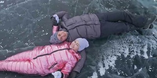 年轻的夫妇有乐趣在冬季散步的背景冰冻结的湖。情侣们躺在有裂缝的透明冰面上，嬉戏、亲吻、拥抱。从以上观点。蜜月。爱情故事。