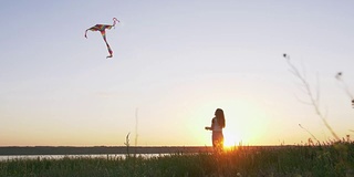 快乐的年轻女子在一个湖泊背景的林间空地上玩风筝在夏季日落