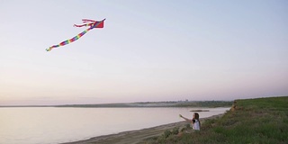 快乐的年轻女子在一个湖泊背景的林间空地上玩风筝在夏季日落