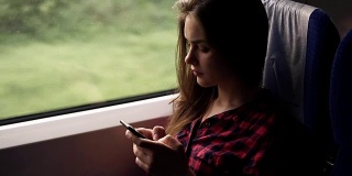 严肃，放松的年轻女孩坐在火车靠窗的地方。乘现代火车旅行。长头发的女孩正在用她的手机滚动或打字。红色格子衬衫。侧视图