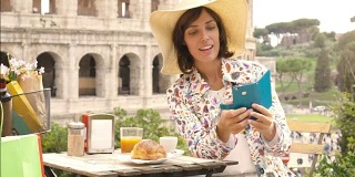 在罗马斗牛场前的一家酒吧餐厅外，一名快乐的年轻游客正在用智能手机自拍，一边喝着咖啡、果汁和玉米片。优雅漂亮的裙子配上大帽子和五颜六色的购物袋