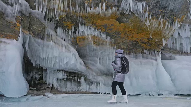 女人在贝加尔湖冰面上的旅行。冬岛之旅。女孩在冰岩石下行走。游客看着美丽的冰洞。极限跋涉和步行。背包客在大自然中休憩。